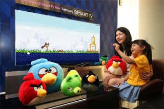 Uma foto de uma criança e seus pais jogando pássaros raivosos em uma tela grande, com pelúcias dos personagens colocados com destaque